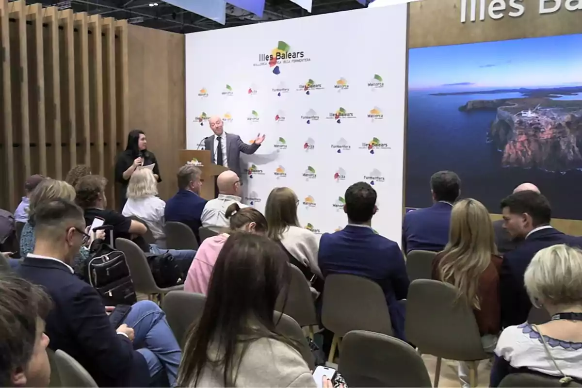 Un grupo de personas asiste a una conferencia en un auditorio moderno mientras un hombre y una mujer presentan en un escenario con el logotipo de Illes Balears y una gran imagen de un paisaje costero al fondo