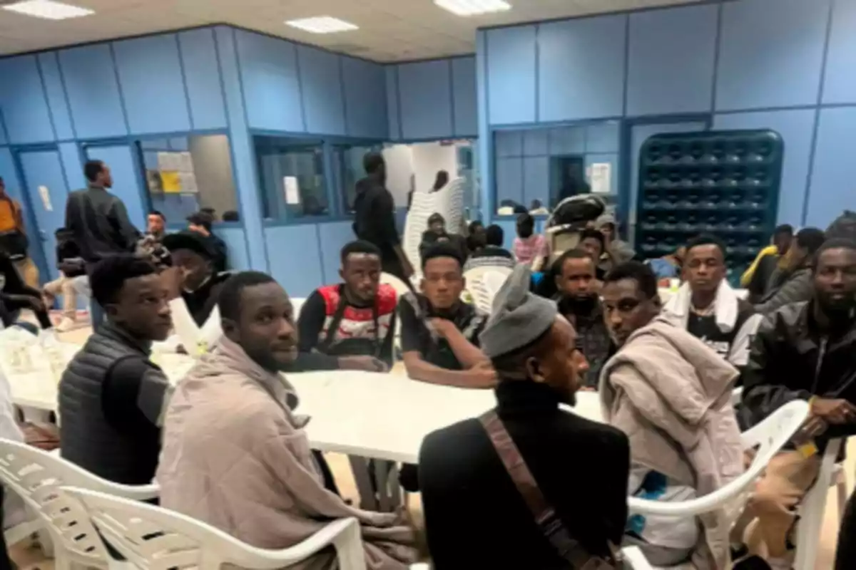 Un grupo de personas sentadas alrededor de una mesa blanca en una sala con paredes azules y sillas de plástico blancas, algunas personas miran hacia la cámara y otras conversan entre sí.