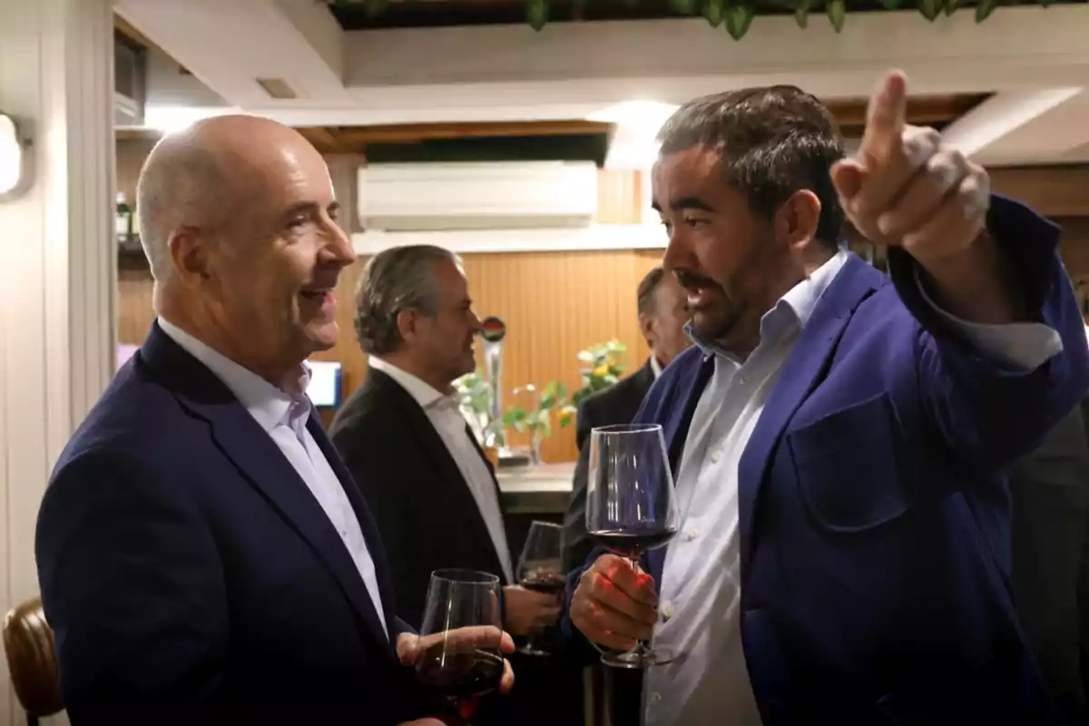 Dos hombres conversan animadamente en un evento social mientras sostienen copas de vino tinto y uno de ellos señala hacia el fondo del lugar