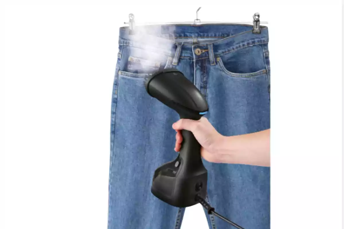 Persona usando un vaporizador de mano para quitar arrugas de unos pantalones de mezclilla colgados en ganchos