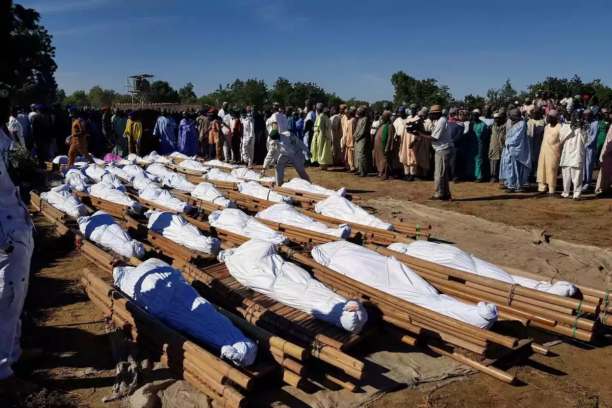 Varias personas envueltas en telas blancas yaciendo sobre camillas de madera mientras un grupo numeroso de personas observa y participa en una ceremonia al aire libre bajo un cielo despejado Varias personas envueltas en telas blancas yaciendo sobre camillas de madera mientras un grupo numeroso de personas observa y participa en una ceremonia al aire libre bajo un cielo despejado