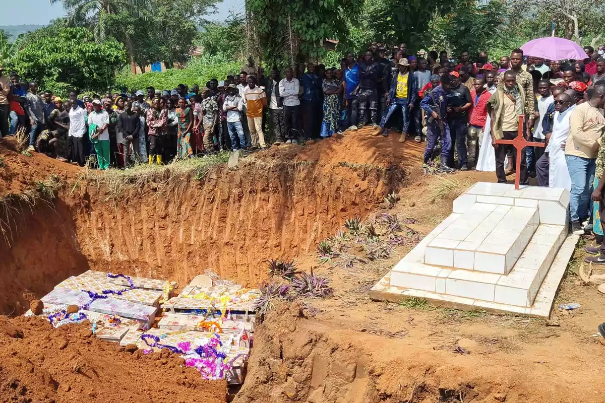 Un grupo numeroso de personas se reúne alrededor de una fosa común donde varios ataúdes decorados con flores están listos para ser enterrados junto a una tumba ya construida en un entorno natural.