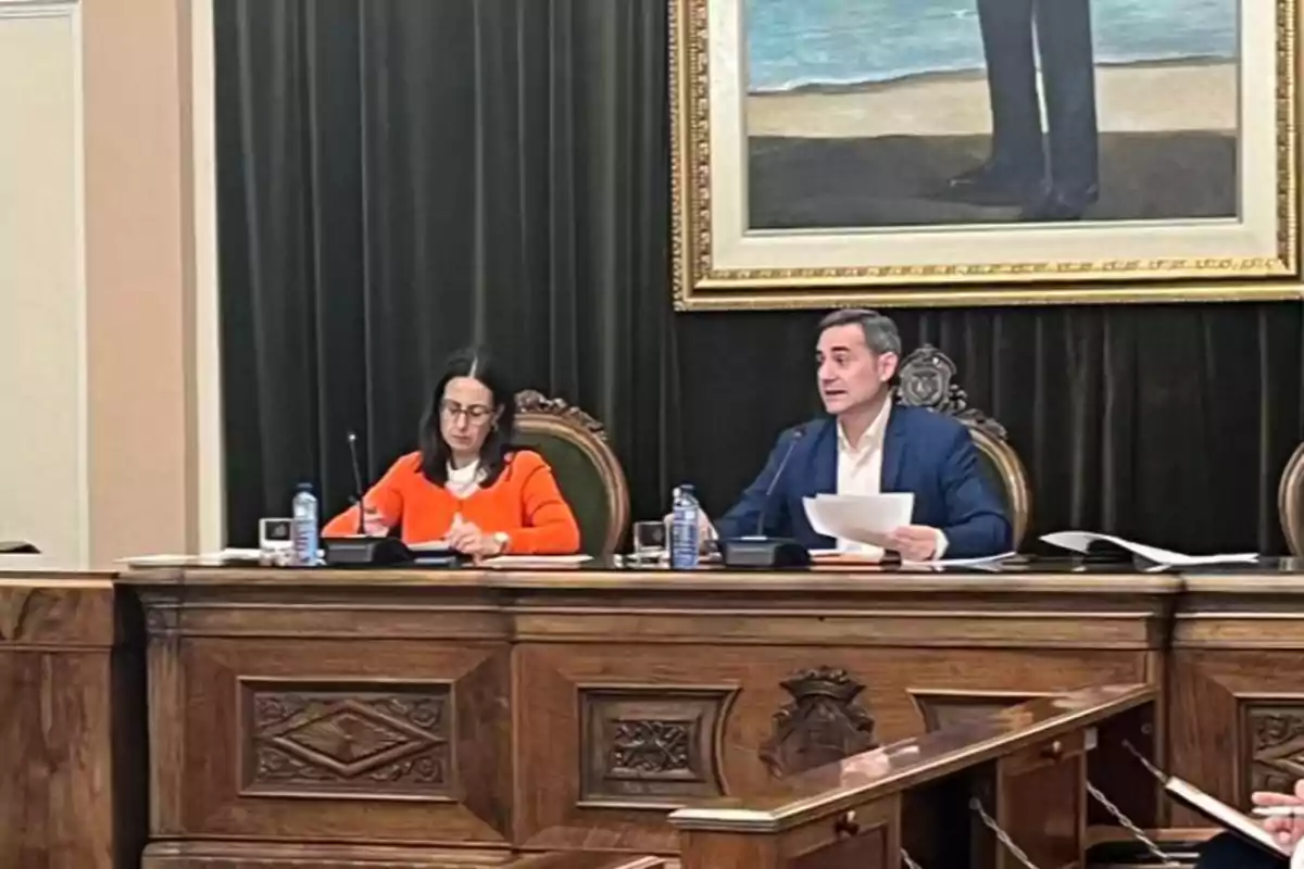 Dos personas sentadas en una mesa de madera en una sala de reuniones, una de ellas con ropa naranja y la otra con saco azul, ambas con papeles y botellas de agua, al fondo una cortina oscura y un cuadro enmarcado