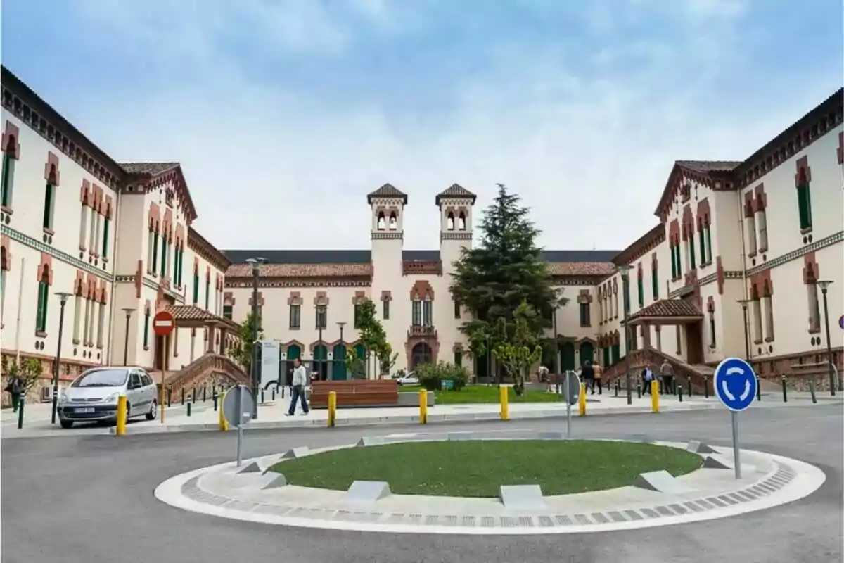 Edificio histórico de fachada blanca con detalles en ladrillo rojo y ventanas verdes, rodeado de una rotonda y algunos árboles, con personas caminando y un coche estacionado