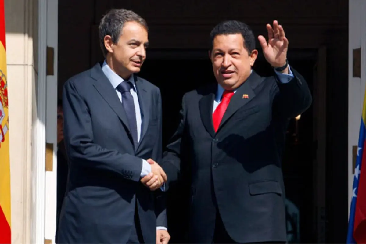 Dos hombres con traje oscuro se dan la mano frente a una puerta mientras uno de ellos saluda levantando la mano y a los lados se ven las banderas de España y Venezuela