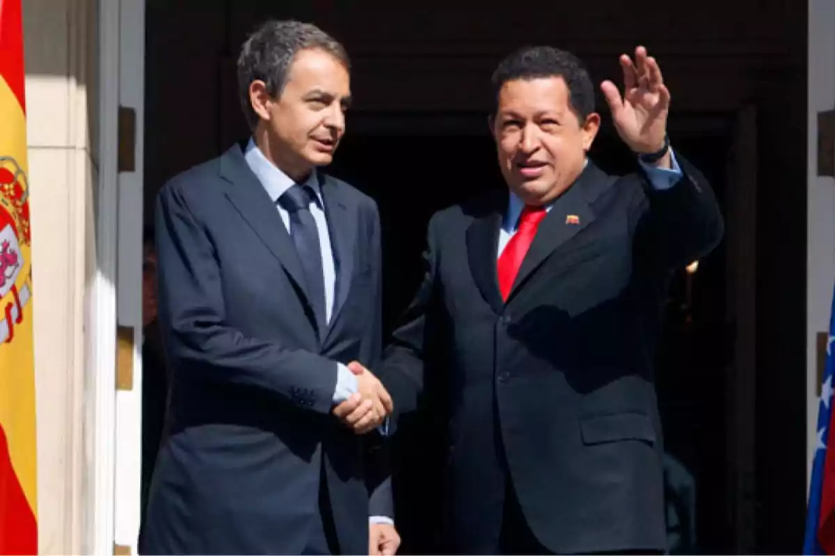 Dos hombres de traje se dan la mano mientras uno de ellos saluda con la mano levantada frente a una bandera de España Dos hombres de traje se dan la mano mientras uno de ellos saluda con la mano levantada frente a una bandera de España