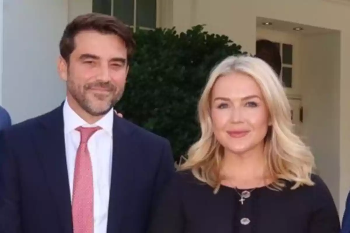 Un hombre con traje oscuro y corbata roja junto a una mujer rubia con cabello suelto y vestido negro posan sonrientes frente a un edificio blanco