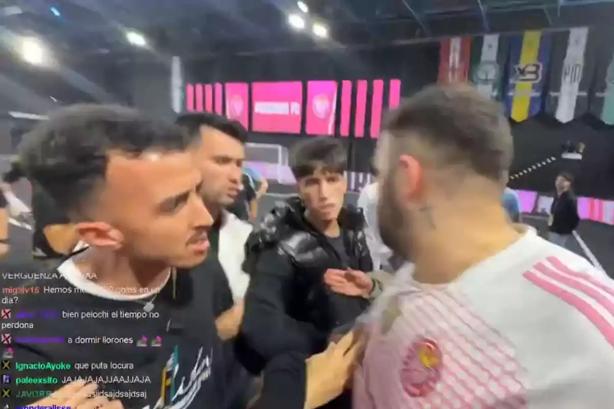 Varios hombres discuten acaloradamente en un evento deportivo bajo techo con pantallas y banderas de fondo.