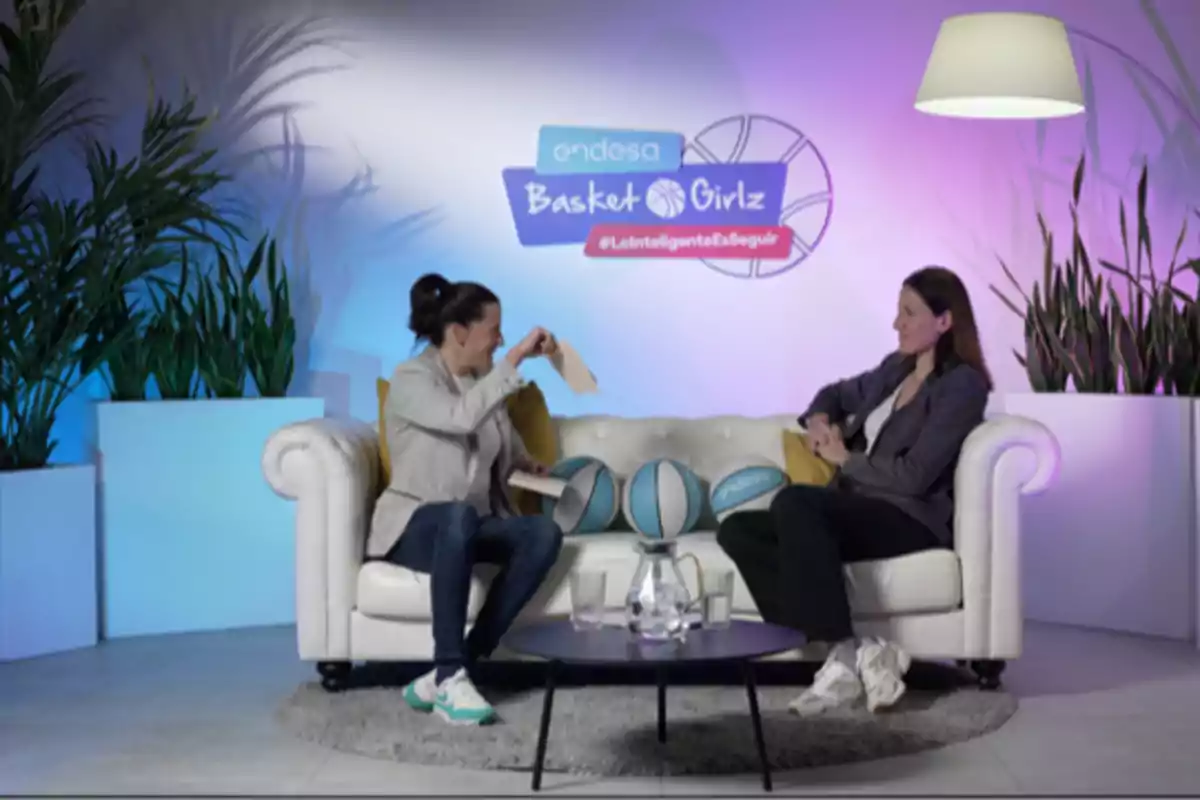 Dos mujeres conversan sentadas en un sofá blanco con balones de baloncesto y plantas decorativas en un set iluminado con luces azules y moradas y un cartel de Basket Girlz al fondo