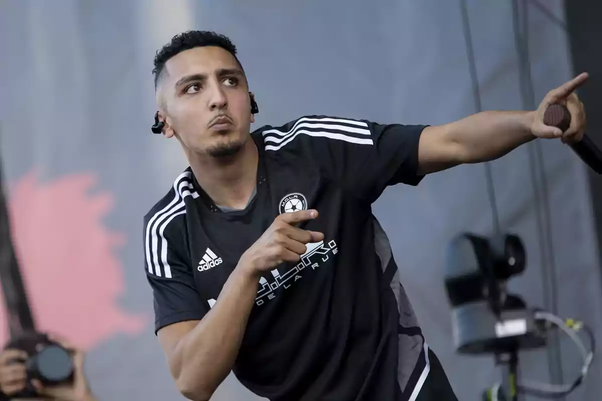 Un hombre con camiseta negra de Adidas sostiene un micrófono y señala hacia un lado mientras está en el escenario.