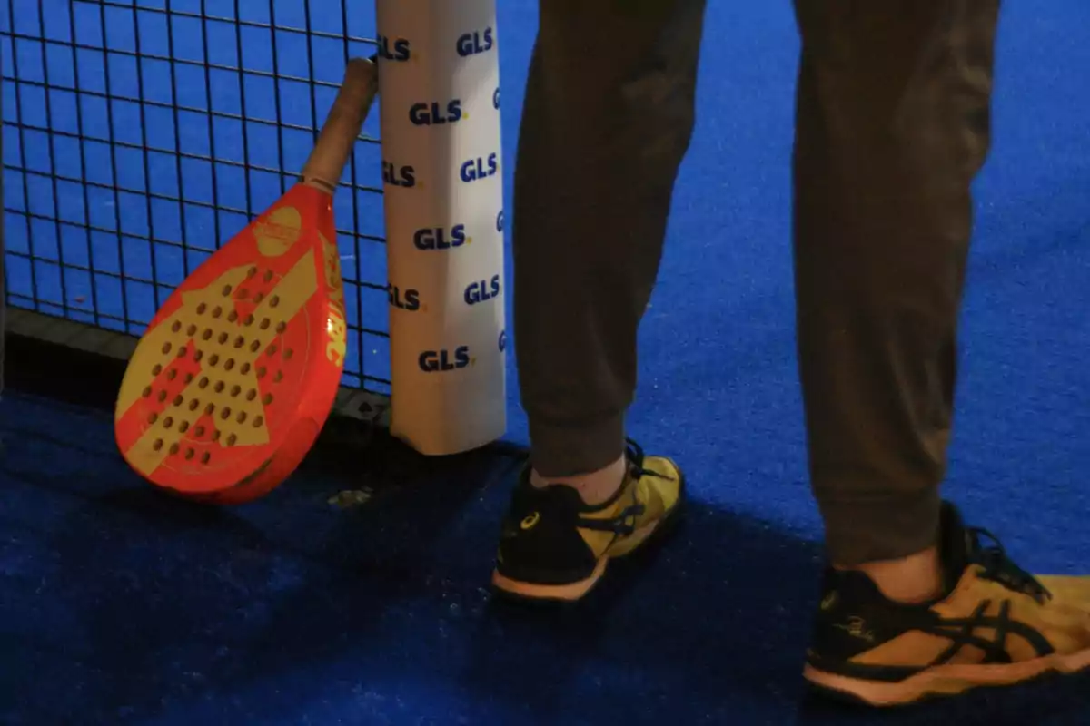 Persona con pantalones deportivos y zapatillas junto a una pala de pádel apoyada en la reja de una cancha con piso azul Persona con pantalones deportivos y zapatillas junto a una pala de pádel apoyada en la reja de una cancha con piso azul