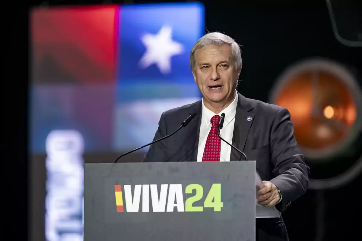 Hombre de cabello canoso y traje oscuro hablando en un podio con el logo VIVA24 y una pantalla de fondo con colores y una estrella blanca.