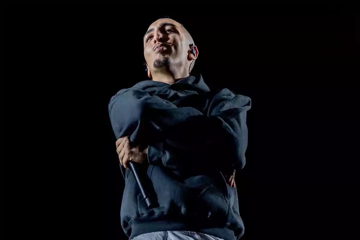 Persona con sudadera oscura y micrófono en la mano, de brazos cruzados y expresión confiada, sobre un fondo negro