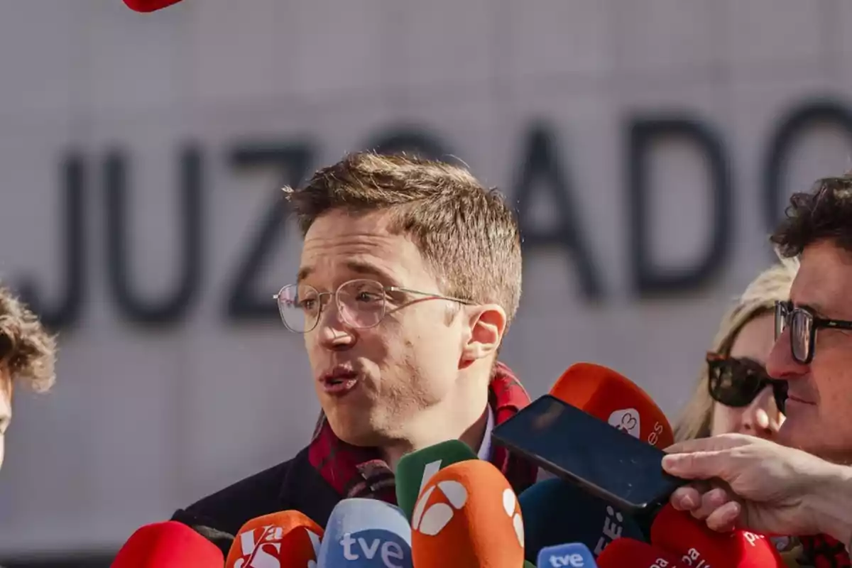 Un hombre con gafas habla frente a varios micrófonos de diferentes medios de comunicación con la palabra juzgado desenfocada al fondo