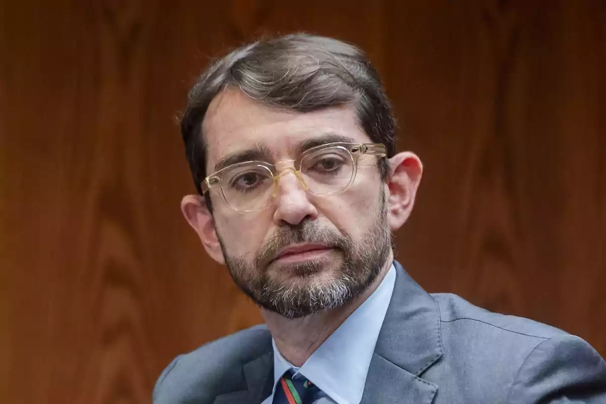 Hombre de cabello corto y barba con gafas transparentes y traje gris sentado frente a una pared de madera Hombre de cabello corto y barba con gafas transparentes y traje gris sentado frente a una pared de madera