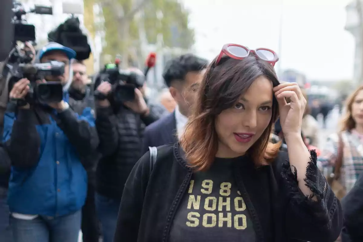 Mujer con gafas de sol en la cabeza y camiseta negra caminando mientras varios camarógrafos la graban en la calle