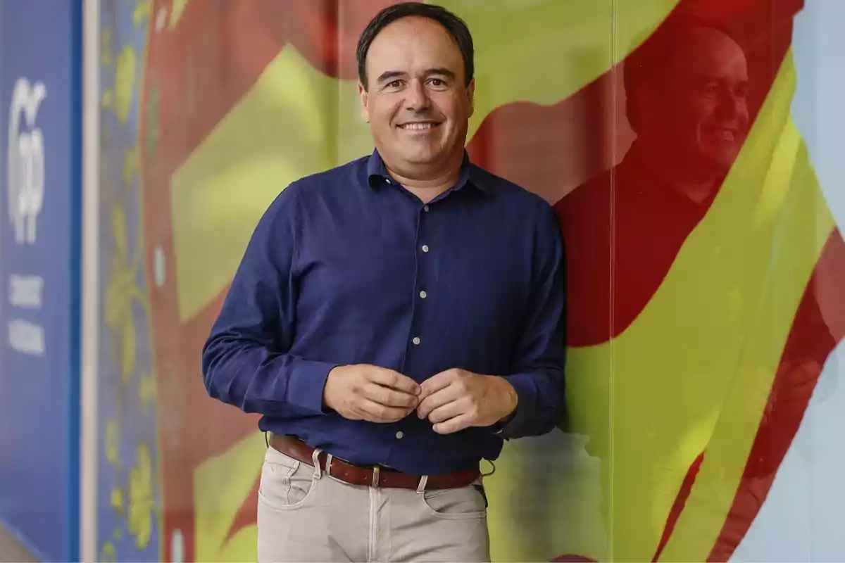 Hombre de camisa azul y pantalón claro sonriendo frente a una pared de colores con su reflejo visible en el vidrio