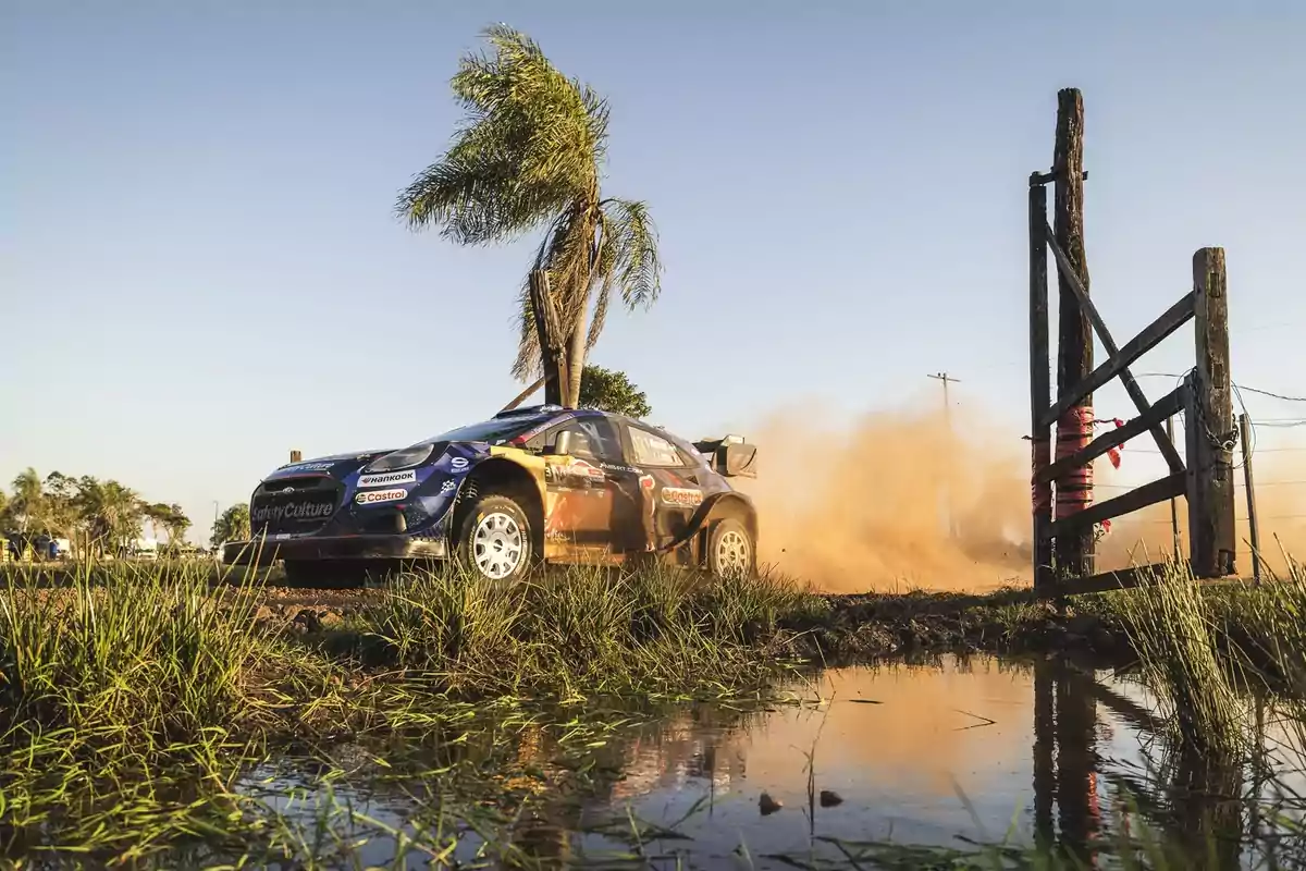 Auto de rally azul avanzando a gran velocidad por un camino de tierra levantando polvo junto a una palmera y un portón de madera reflejados en un charco de agua