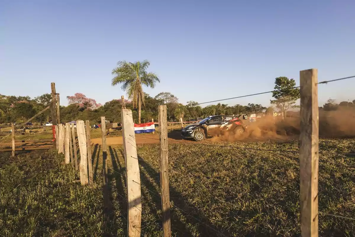 Auto de rally levantando polvo en un camino de tierra rural rodeado de postes de madera y una bandera de Paraguay, con árboles y cielo despejado al fondo