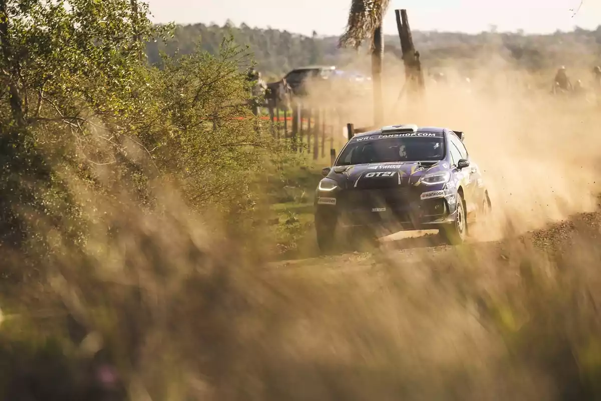 Auto de rally avanzando a gran velocidad por un camino de tierra rodeado de vegetación y levantando una nube de polvo