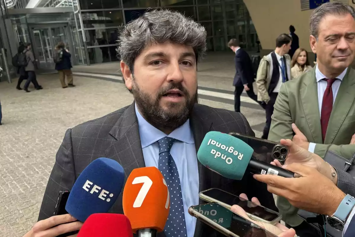 Un hombre con barba y traje oscuro responde preguntas de varios periodistas que sostienen micrófonos de diferentes medios frente a un edificio moderno