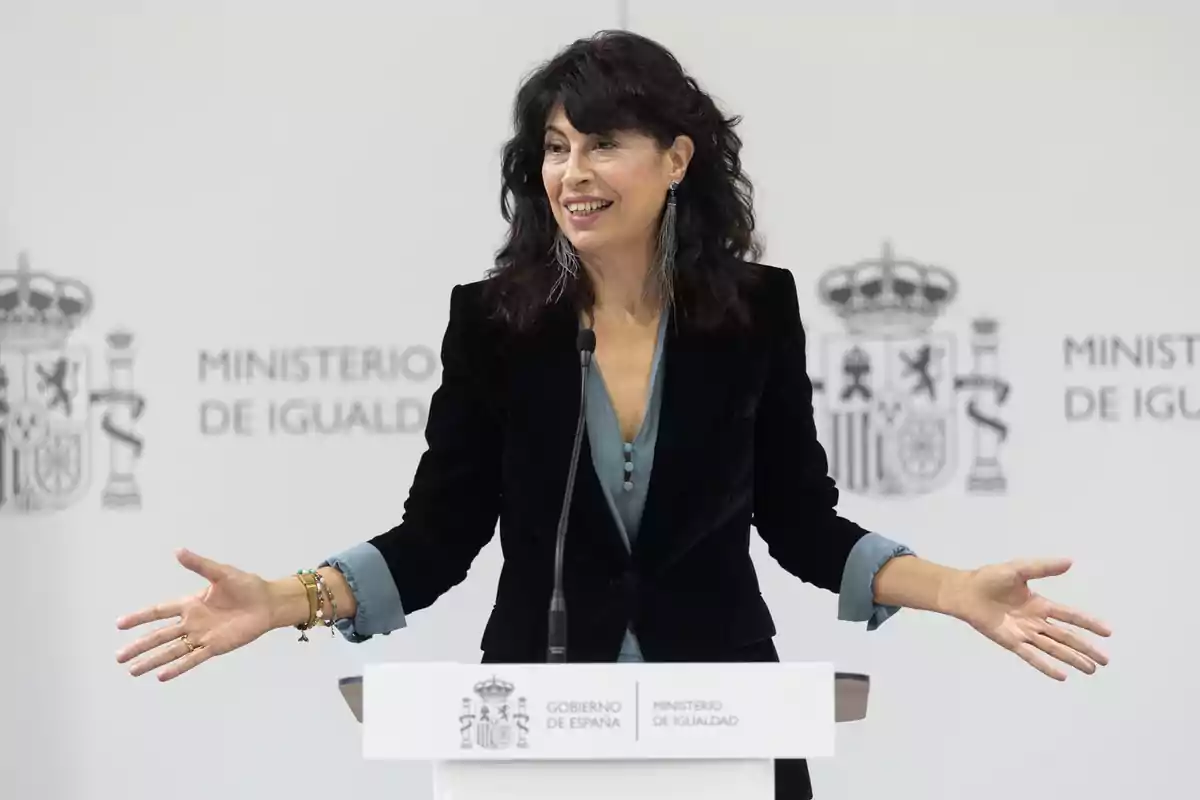 Mujer de cabello oscuro y suelto hablando sonriente en un atril del Ministerio de Igualdad de España con los brazos abiertos Mujer de cabello oscuro y suelto hablando sonriente en un atril del Ministerio de Igualdad de España con los brazos abiertos