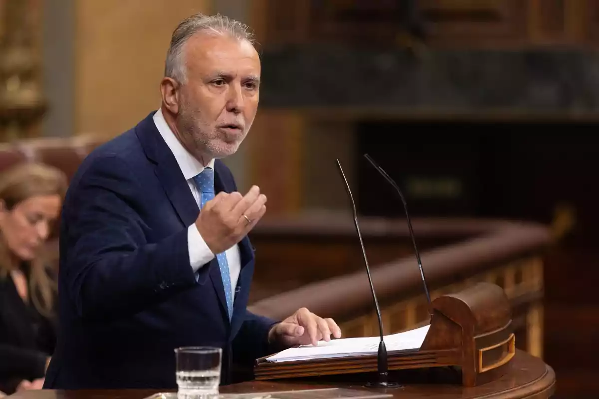 Hombre de cabello canoso y traje azul hablando en un podio de madera con micrófonos en un entorno parlamentario