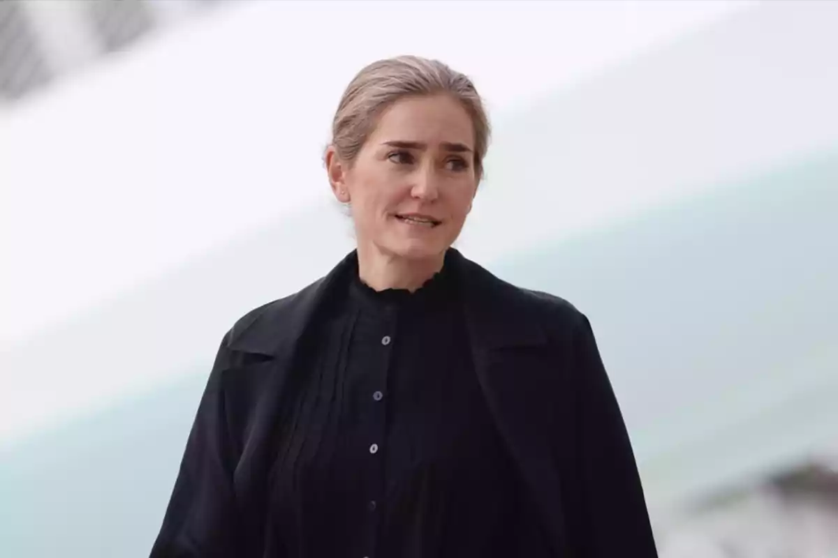 Mujer de cabello rubio recogido vestida con ropa oscura mirando hacia un lado con expresión seria