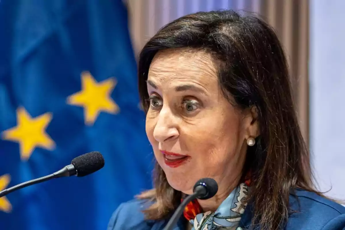Mujer de cabello oscuro hablando frente a un micrófono con una bandera azul con estrellas amarillas de fondo