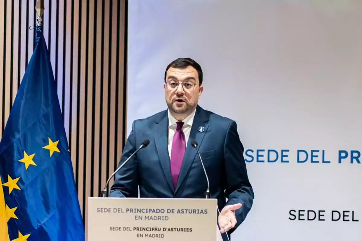 Hombre con gafas y traje azul hablando en un atril con micrófonos junto a una bandera azul con estrellas amarillas y un cartel que dice sede del Principado de Asturias en Madrid Hombre con gafas y traje azul hablando en un atril con micrófonos junto a una bandera azul con estrellas amarillas y un cartel que dice sede del Principado de Asturias en Madrid