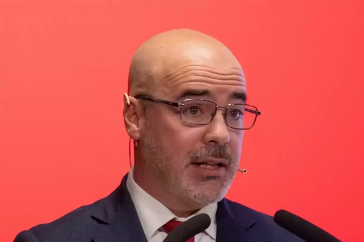 Hombre calvo con gafas y barba hablando frente a un micrófono con fondo rojo