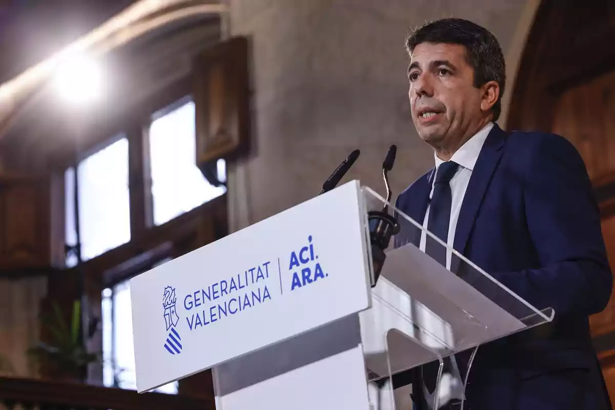 Hombre con traje azul hablando en un atril de la Generalitat Valenciana en un interior con ventanas al fondo Hombre con traje azul hablando en un atril de la Generalitat Valenciana en un interior con ventanas al fondo
