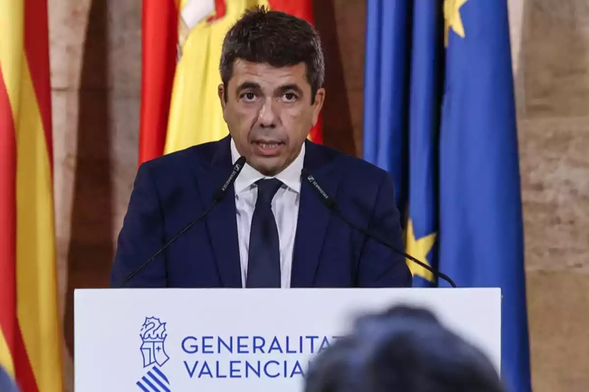 Hombre con traje azul hablando en un atril de la Generalitat Valenciana con banderas de fondo
