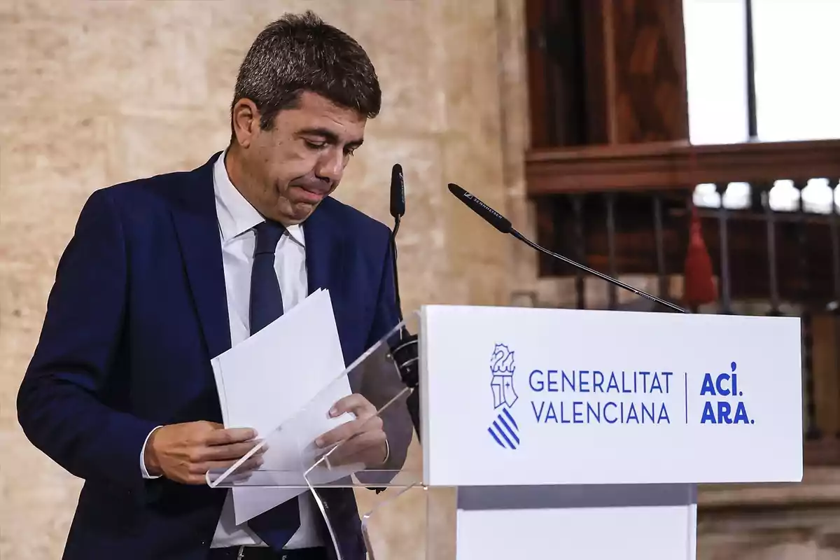 Hombre con traje azul sosteniendo papeles frente a un atril de la Generalitat Valenciana durante un evento oficial Hombre con traje azul sosteniendo papeles frente a un atril de la Generalitat Valenciana durante un evento oficial
