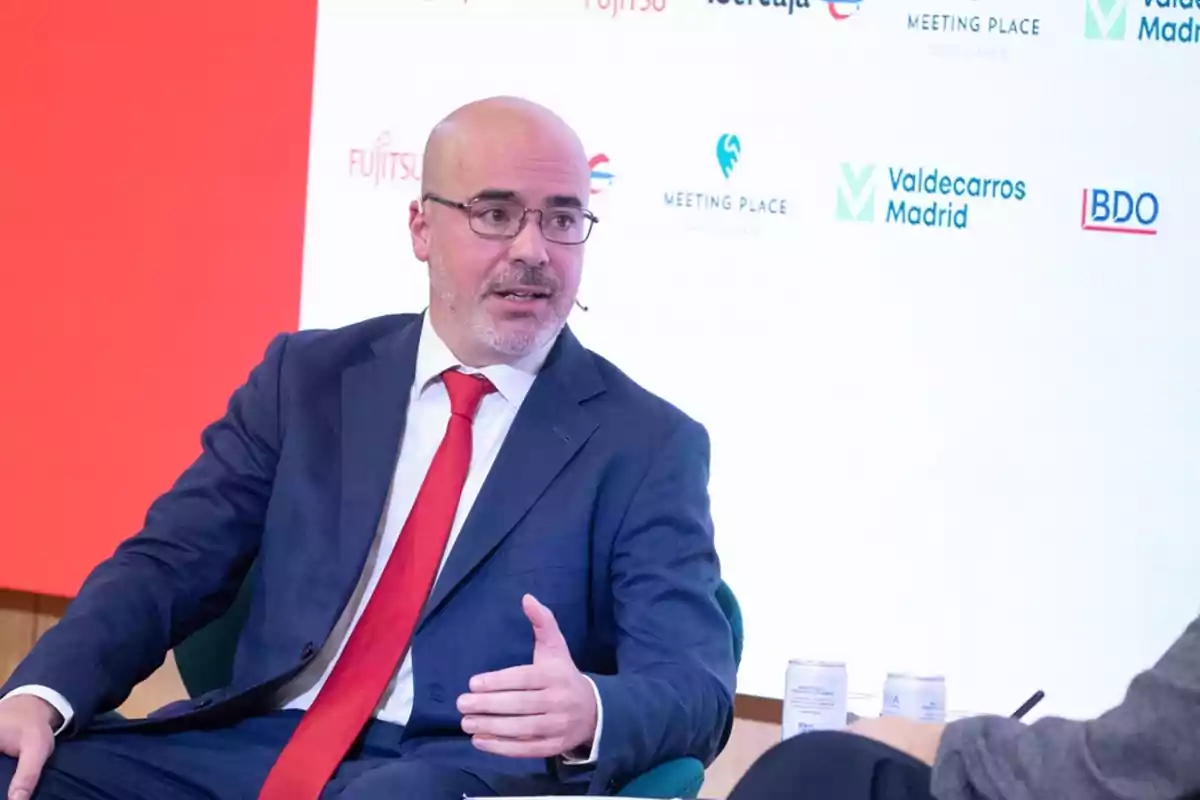 Hombre calvo con gafas y traje azul hablando durante un evento empresarial con fondo de logotipos de empresas. Hombre calvo con gafas y traje azul hablando durante un evento empresarial con fondo de logotipos de empresas.