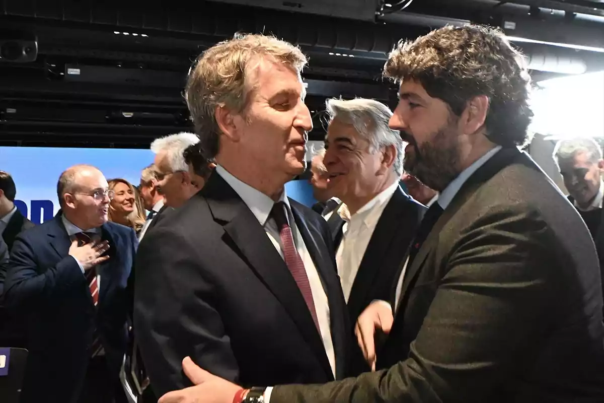 Dos hombres de traje se saludan sonrientes en un evento rodeados de otras personas en un ambiente profesional. Dos hombres de traje se saludan sonrientes en un evento rodeados de otras personas en un ambiente profesional.