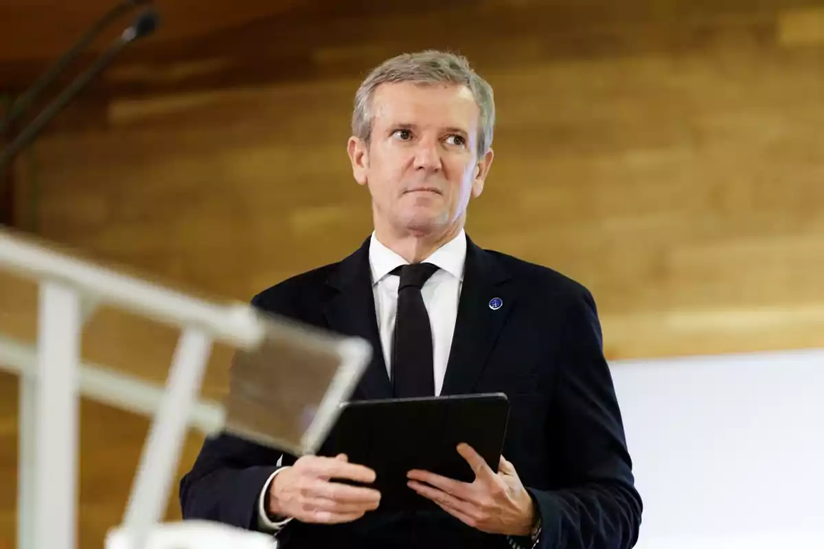 Hombre de cabello canoso con traje oscuro y corbata negra sosteniendo una tableta en un entorno interior con fondo de madera