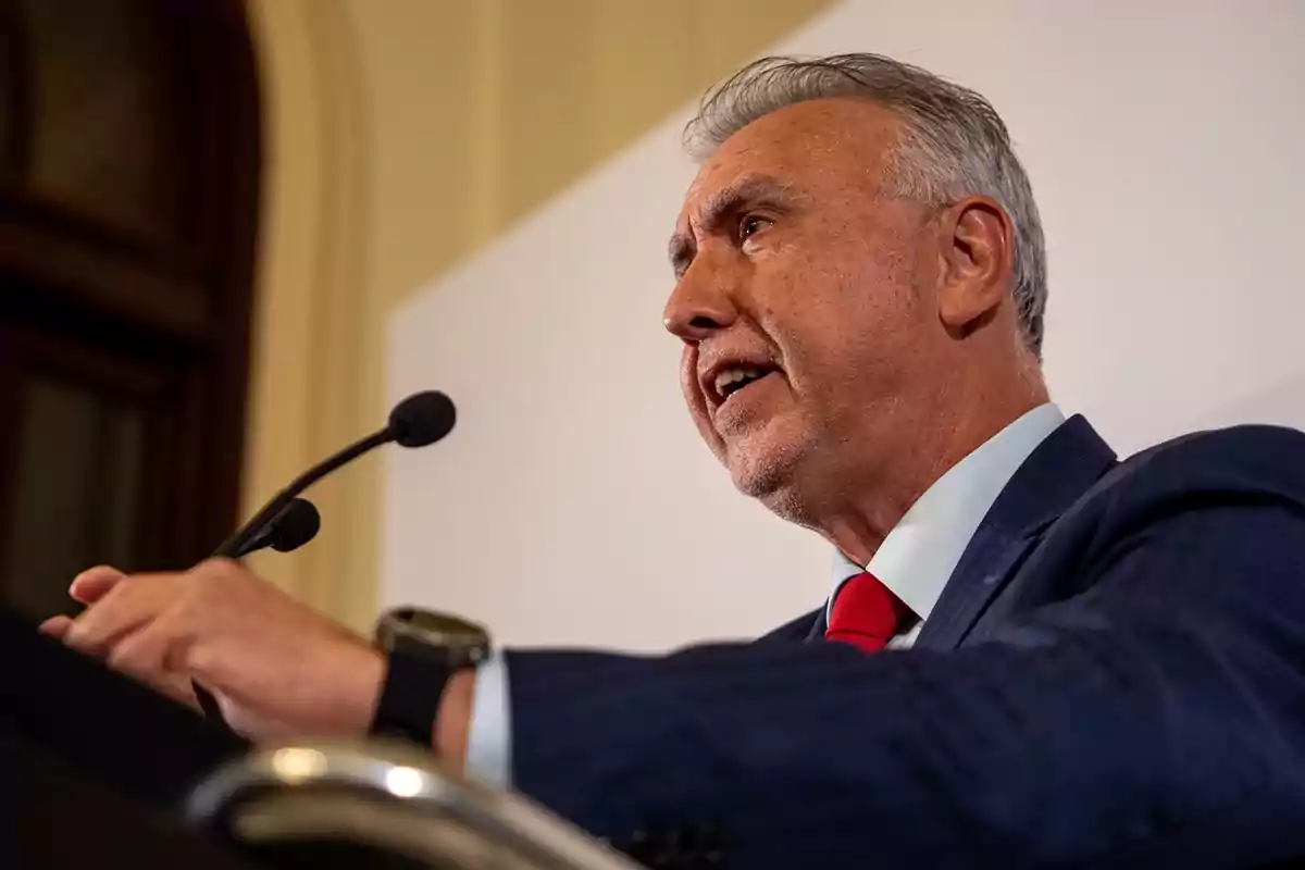Hombre de cabello canoso con traje azul y corbata roja hablando frente a un micrófono en un podio