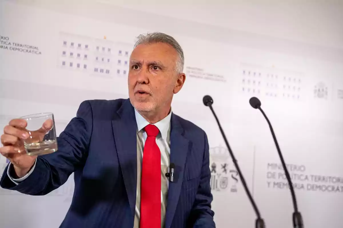 Hombre de traje azul y corbata roja sostiene un vaso de agua mientras habla frente a dos micrófonos en un podio institucional