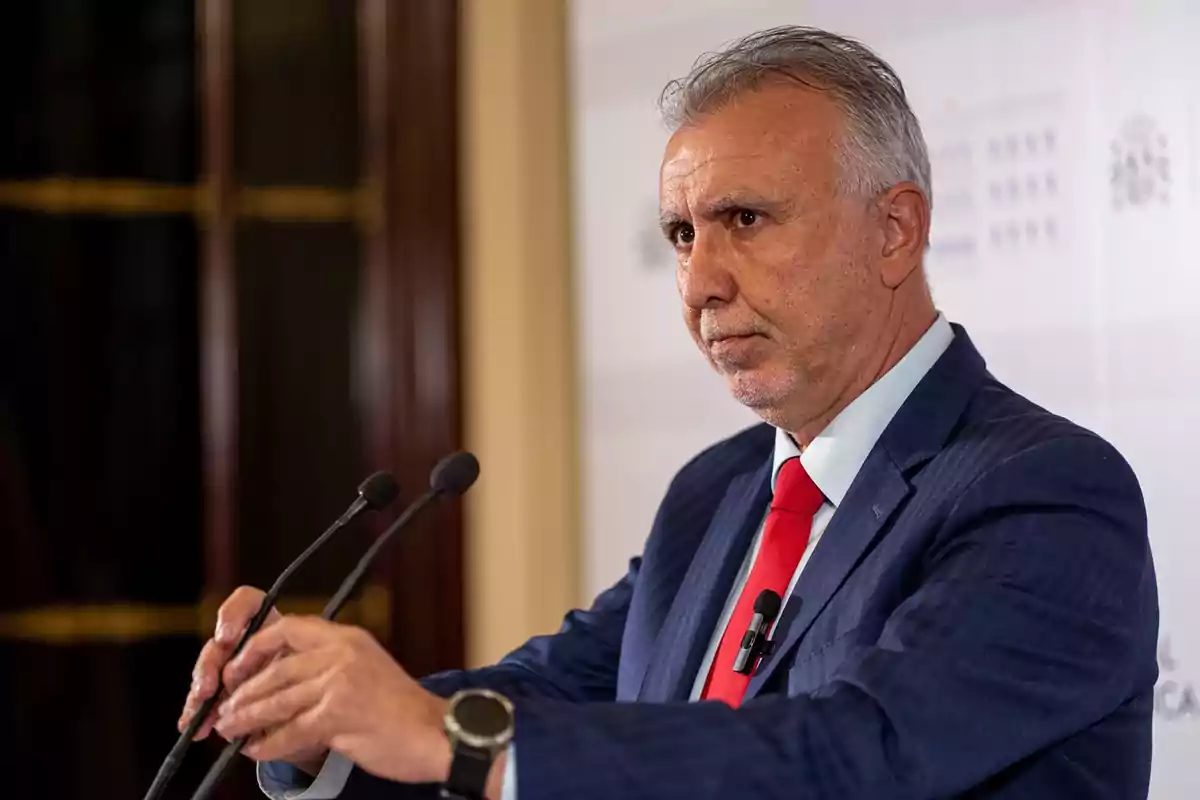 Hombre de cabello canoso con traje azul y corbata roja hablando frente a dos micrófonos en un entorno formal Hombre de cabello canoso con traje azul y corbata roja hablando frente a dos micrófonos en un entorno formal