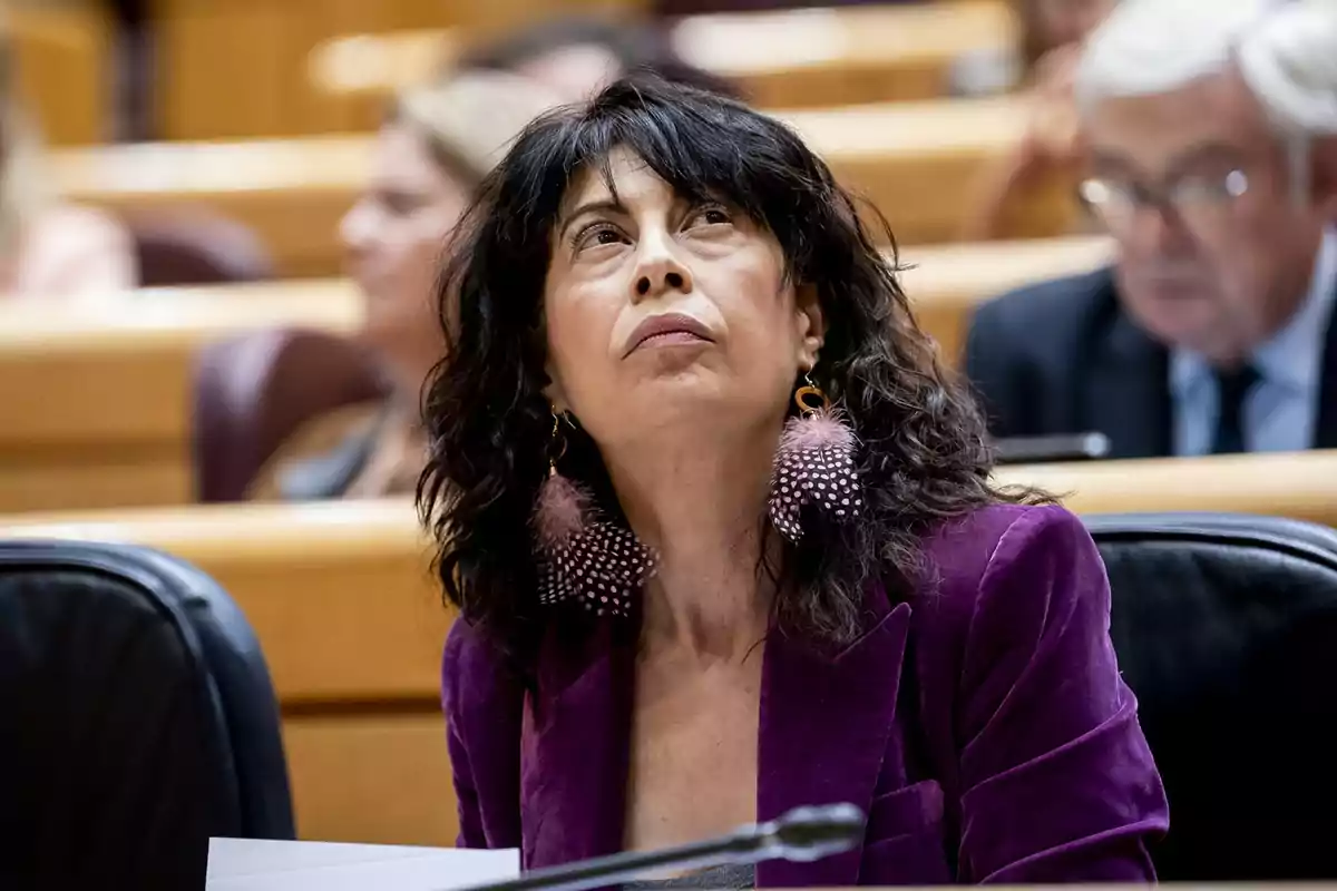 Mujer de cabello oscuro y rizado con chaqueta morada y pendientes grandes sentada en una sala de conferencias mirando hacia arriba
