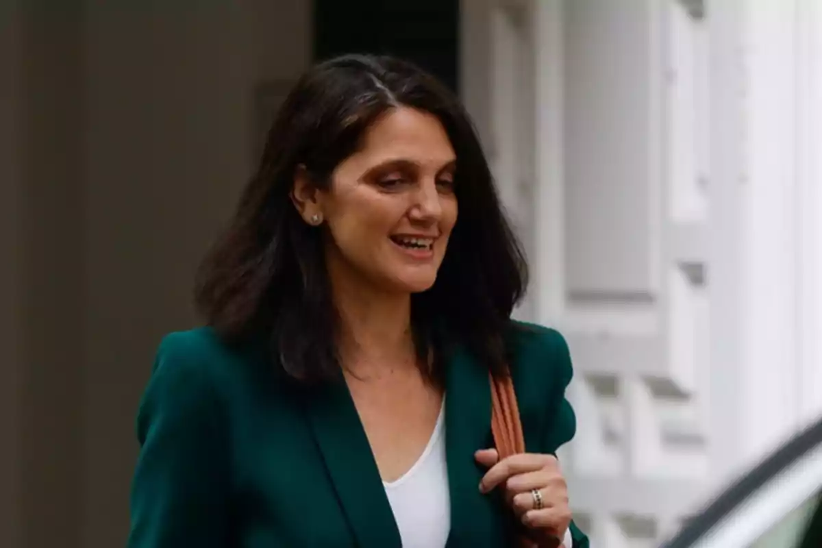 Mujer de cabello oscuro y suelto viste un saco verde y lleva una bolsa al hombro mientras sonríe