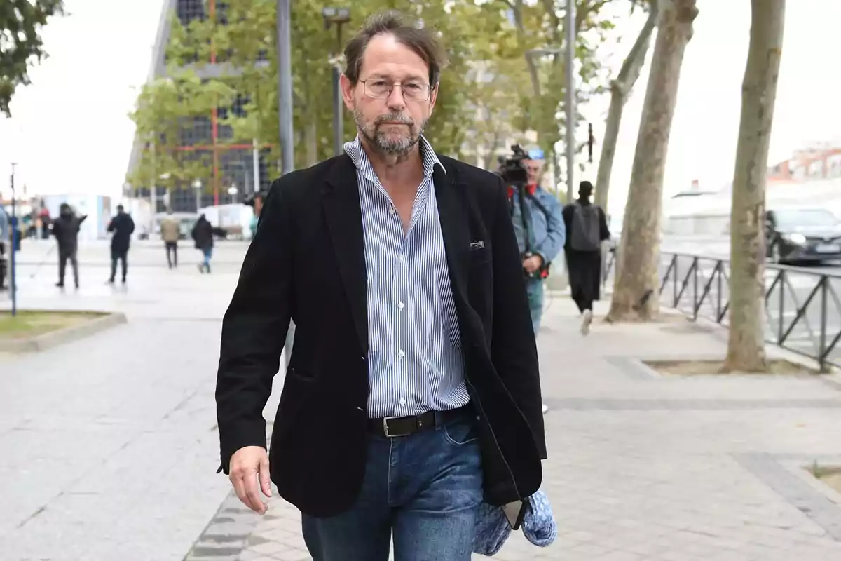 Hombre de mediana edad con barba y gafas caminando por una acera en una ciudad con árboles y personas al fondo