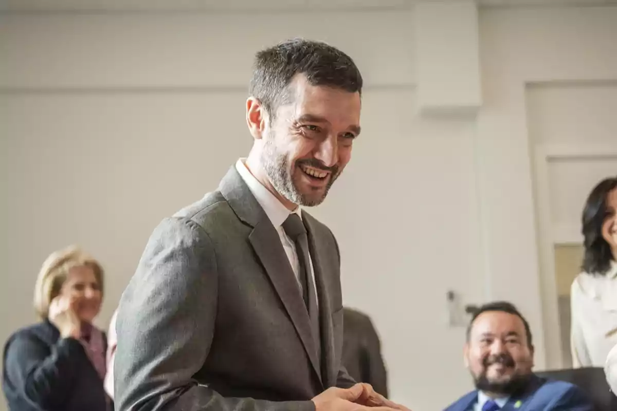Hombre con barba y traje gris sonriendo en una reunión con otras personas al fondo Hombre con barba y traje gris sonriendo en una reunión con otras personas al fondo
