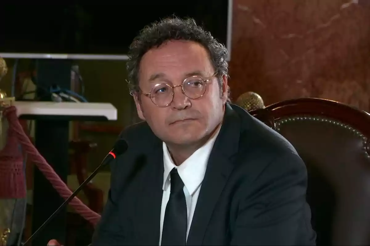 Hombre de cabello rizado y gafas con traje oscuro y corbata negra sentado frente a un micrófono en un entorno formal