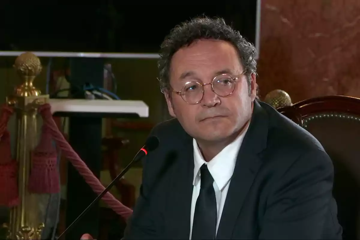 Hombre de mediana edad con gafas y traje oscuro sentado frente a un micrófono en un entorno formal