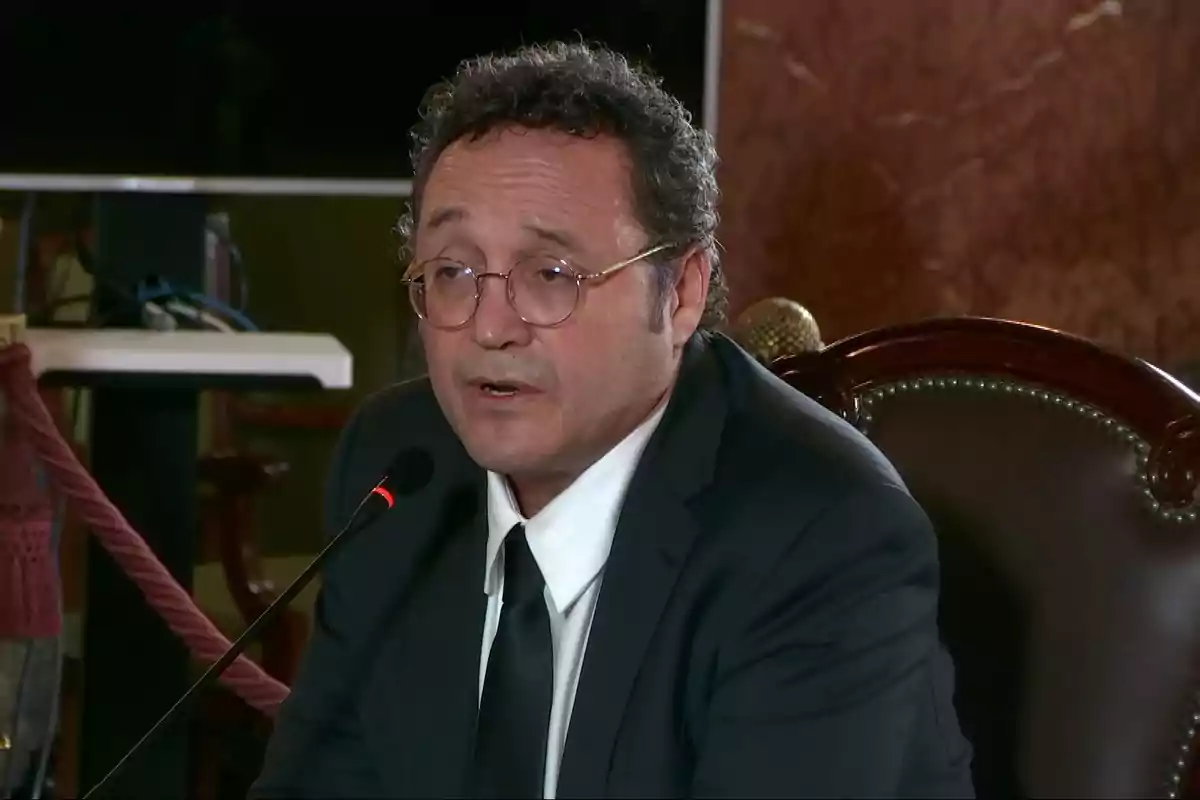 Hombre de cabello rizado y gafas hablando frente a un micrófono mientras viste traje y corbata en un entorno formal Hombre de cabello rizado y gafas hablando frente a un micrófono mientras viste traje y corbata en un entorno formal