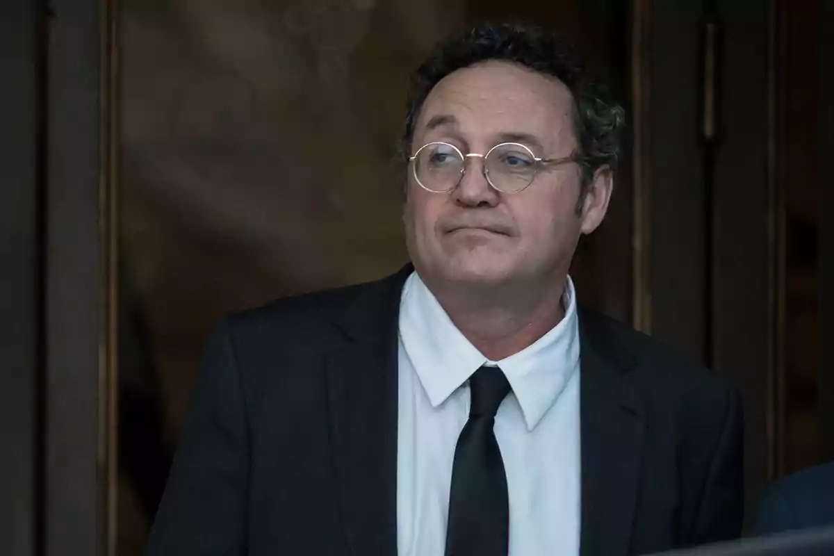 Hombre de cabello rizado y gafas con traje negro y corbata, de pie frente a una puerta de madera Hombre de cabello rizado y gafas con traje negro y corbata, de pie frente a una puerta de madera