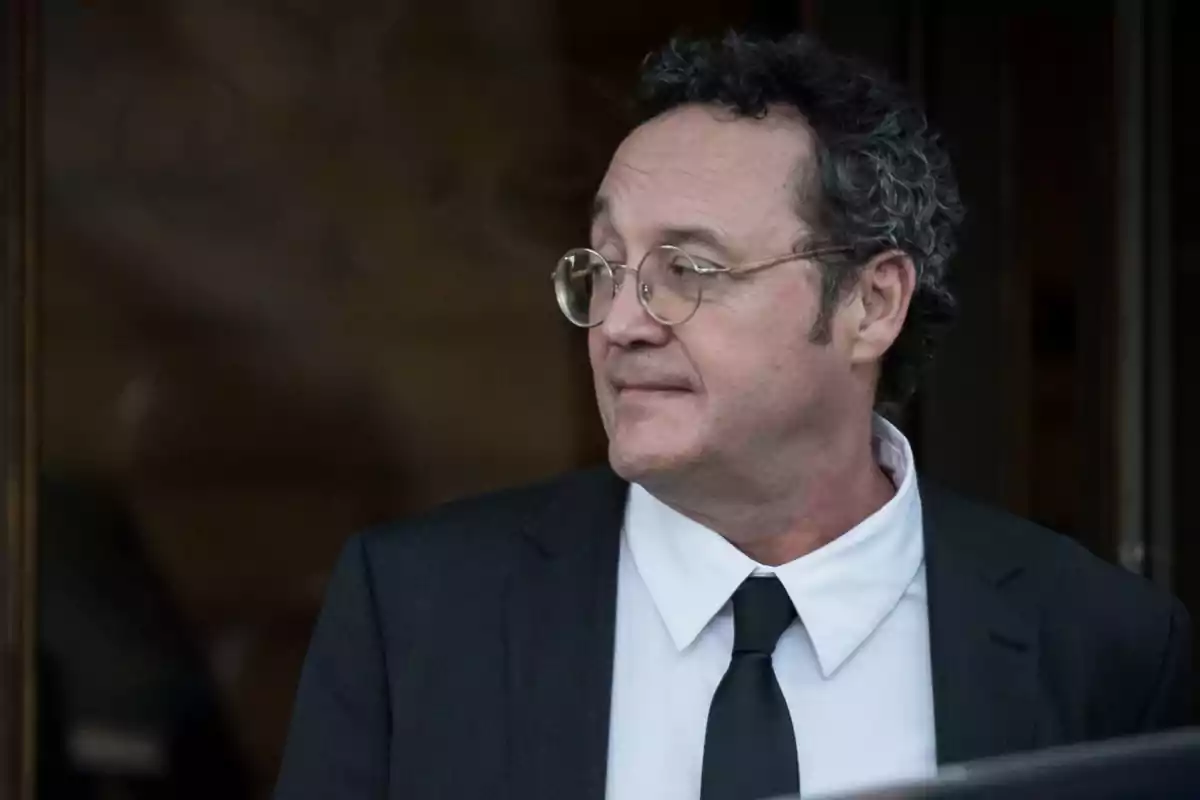 Hombre de cabello rizado y gafas con traje negro y corbata mirando hacia un lado