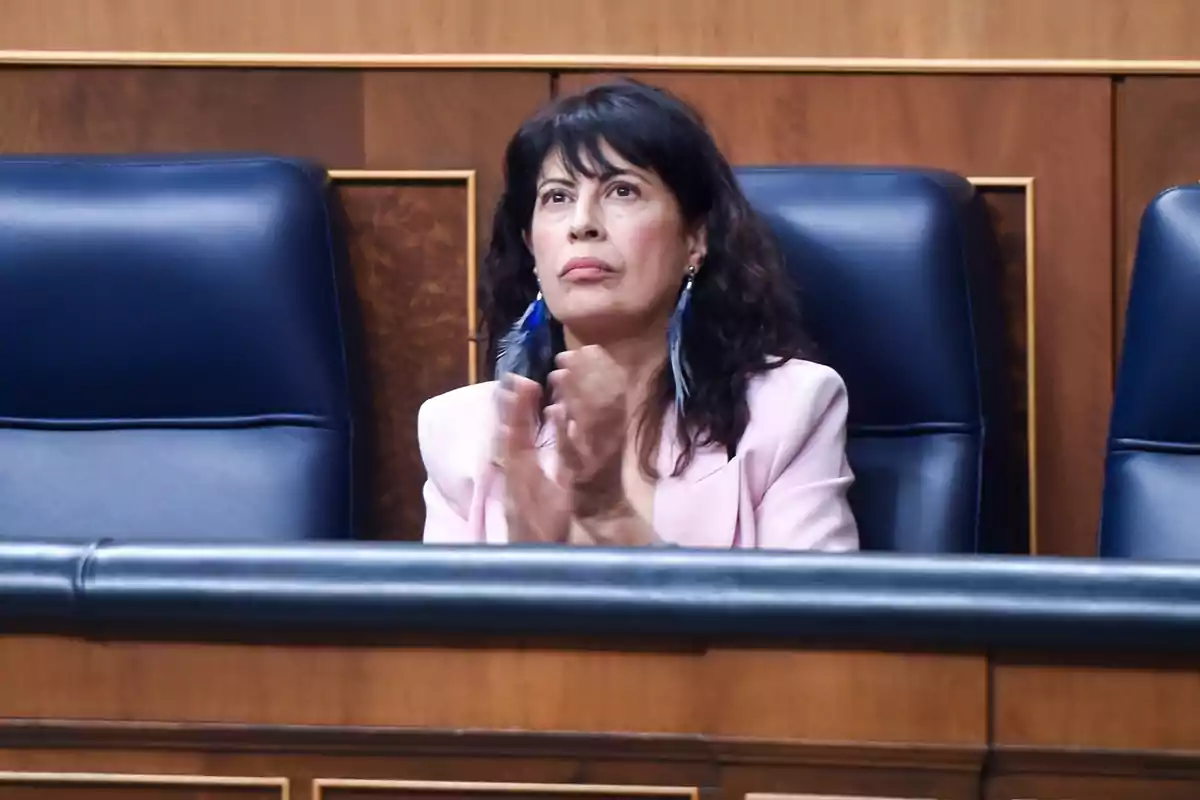 Mujer de cabello oscuro sentada en una silla azul aplaudiendo y vistiendo un saco rosa claro Mujer de cabello oscuro sentada en una silla azul aplaudiendo y vistiendo un saco rosa claro